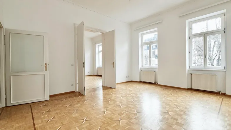 Helles, leeres Zimmer mit Fischgrätparkett, hohen Decken und großen Fenstern, bereit zur individuellen Gestaltung.