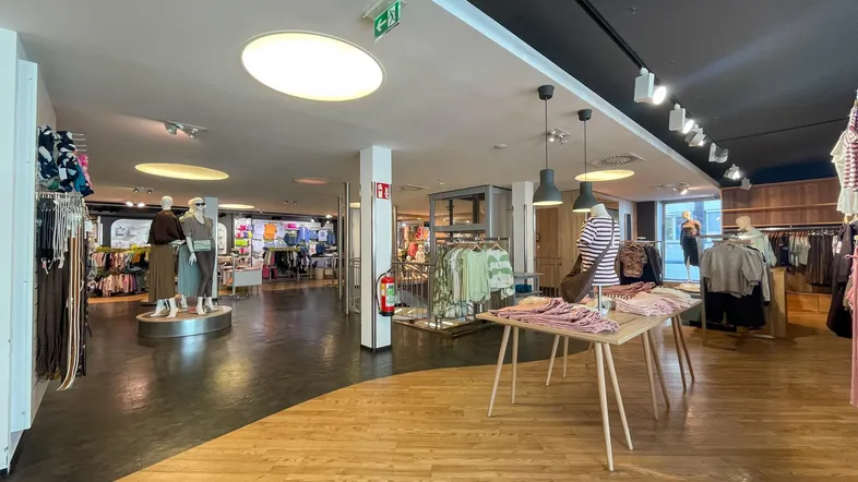 Heller und geräumiger Verkaufsraum mit Holzboden und moderner Beleuchtung, präsentiert Kleidung auf Tischen und Mannequins.