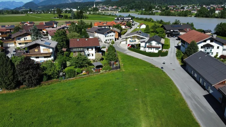 Luftaufnahme einer Wohnsiedlung mit grünen Feldern, Fluss und majestätischen Bergen im Hintergrund.