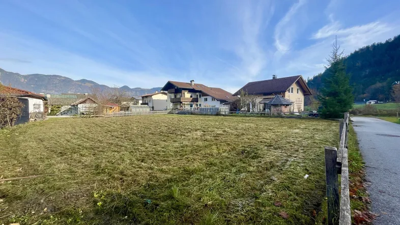 Grüne Wiese mit umliegenden Häusern und Bergpanorama im Hintergrund unter blauem Himmel.
