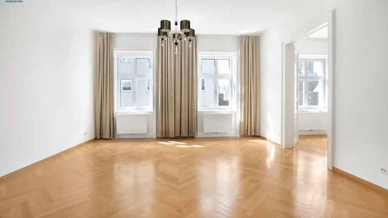 Lichtdurchflutetes Wohnzimmer mit elegantem Fischgrätparkett und zwei hohen Fenstern mit Vorhängen.