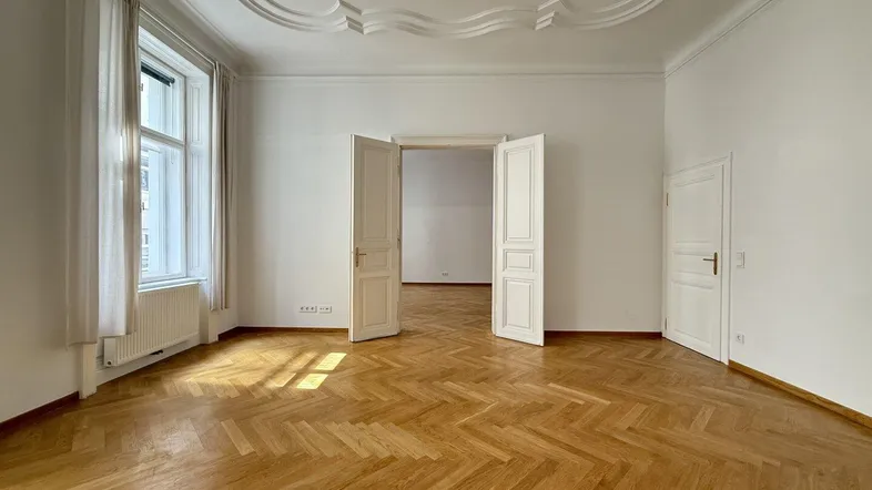 Geräumiges Wohnzimmer mit Fischgrätparkett, hohen Decken und Stuckverzierungen, durch Doppeltüren verbunden.
