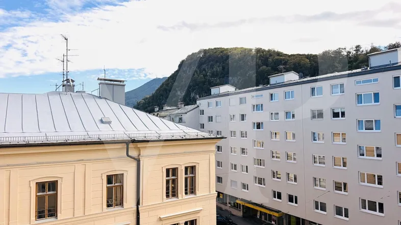 Blick aus dem Fenster auf umliegende Gebäude und bewaldete Berge im Hintergrund unter blauem Himmel.