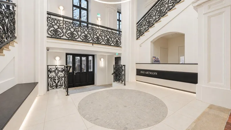 Großzügige Eingangshalle mit eleganter Treppe, schmiedeeisernem Geländer und Rezeption.
