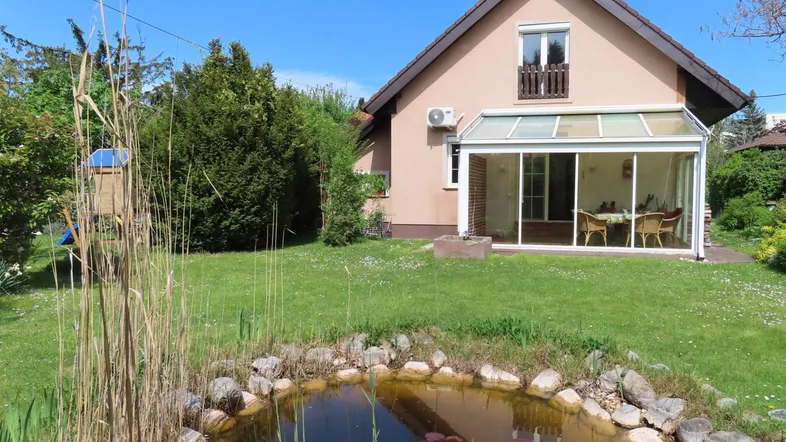 Idyllisches Einfamilienhaus mit Wintergarten, großem Garten und einem kleinen Teich unter blauem Himmel.