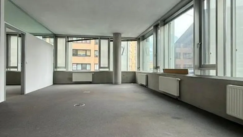 Geräumiges, leeres Büro mit großen Fenstern, grauem Teppichboden und Heizkörpern entlang der Wand.