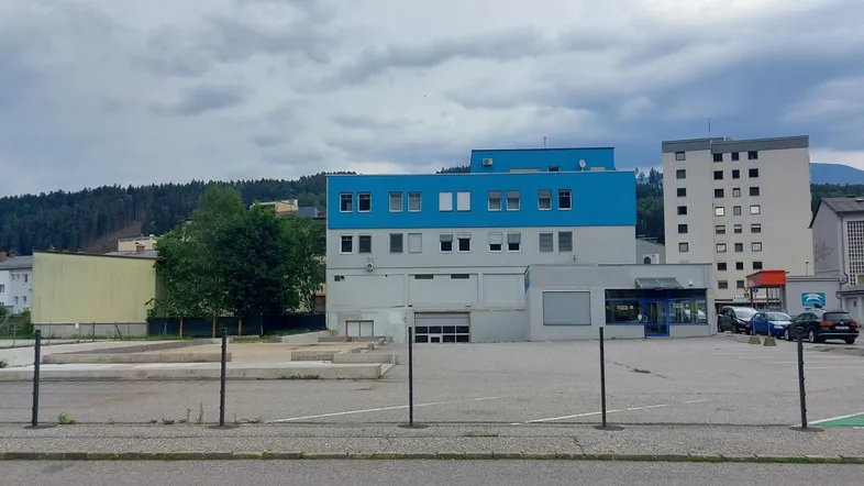 Gebäude mit blauer und grauer Fassade, umgeben von einem Parkplatz und weiteren Gebäuden in einer bergigen Landschaft.