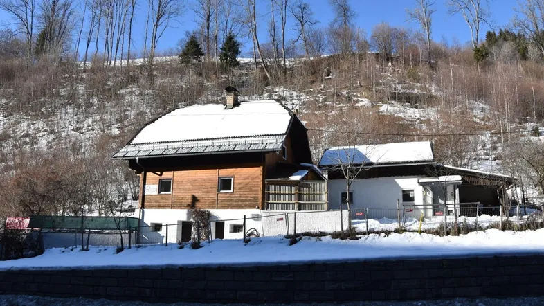 Traditionelles Holzhaus mit schneebedecktem Dach in einer winterlichen Berglandschaft, umgeben von Bäumen.