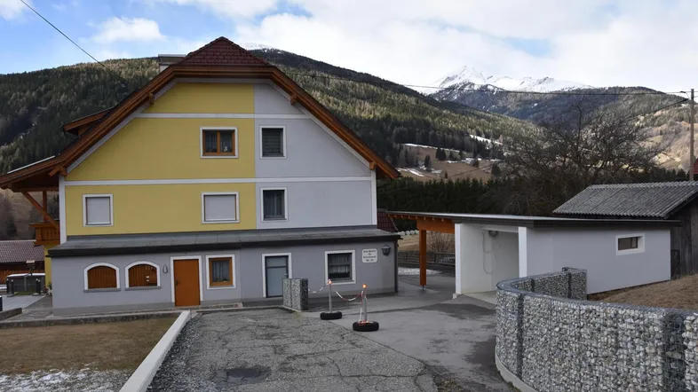 Hausfassade mit gelben und grauen Putzflächen, umgeben von einer Berglandschaft und Carport.