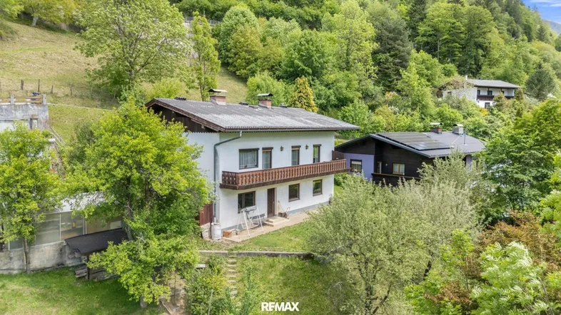 Traditionelles Haus mit Balkon und Satteldach, umgeben von grünen Hügeln und Bäumen.