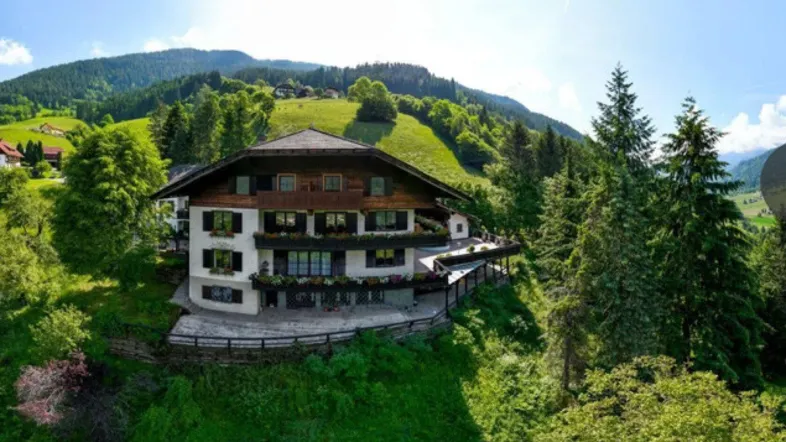 Traditionelles Alpenhaus in idyllischer Hanglage mit mehreren Balkonen und Terrassen, umgeben von grünen Hügeln.