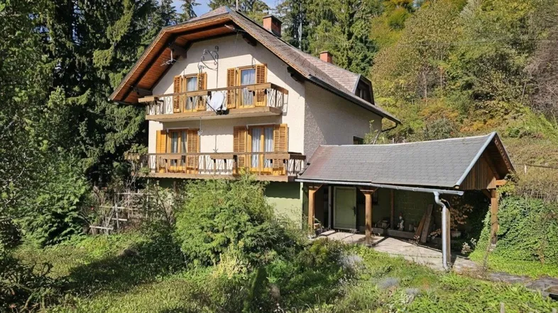 Traditionelles Haus mit Holzbalkonen und Carport, umgeben von üppiger Natur und hohen Bäumen.