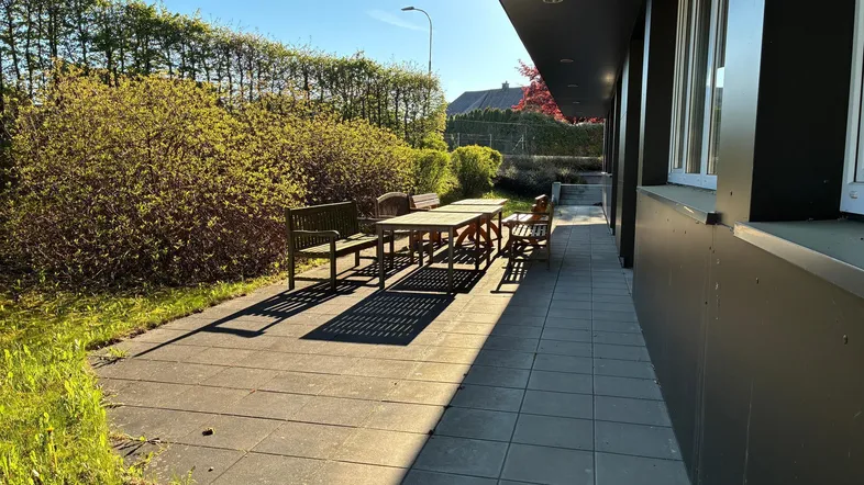 Geräumige Außenterrasse mit Holztisch und Bänken, umgeben von grünen Büschen unter strahlendem Sonnenschein.