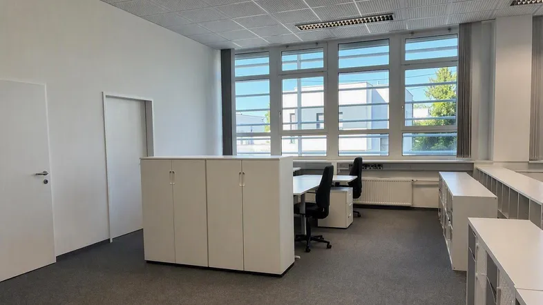 Helles Großraumbüro mit mehreren Arbeitsplätzen, weißen Schränken und großen Fenstern für viel Tageslicht.