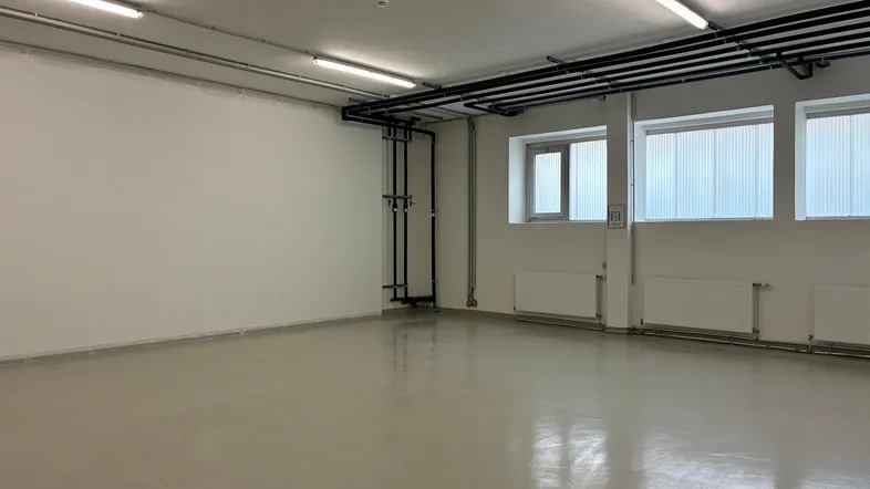 Großer, heller Raum mit weißen Wänden, grauem Boden und sichtbaren Installationen an Decke und Wand.