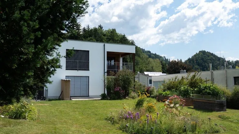 Modernes Haus mit weißer Fassade und gepflegtem Gartenbereich vor grüner Bergkulisse.