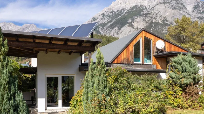 Hausfassade mit Photovoltaikanlage, Terrasse und beeindruckendem Bergblick unter blauem Himmel.