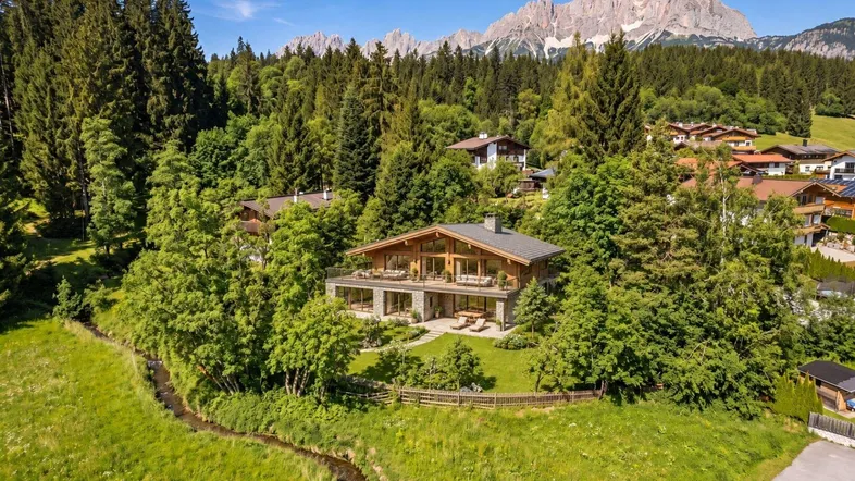Luxuriöses Holzhaus mit Steinfassade, umgeben von grüner Natur und majestätischen Bergen im Hintergrund.