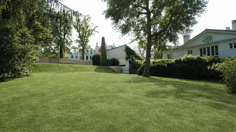 Großer, gepflegter Rasenbereich mit altem Baumbestand und eleganten Gebäuden im Hintergrund unter blauem Himmel.