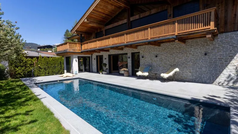 Modernes Chalet mit Außenpool, Steinfassade und Holzbalkonen unter blauem Himmel.