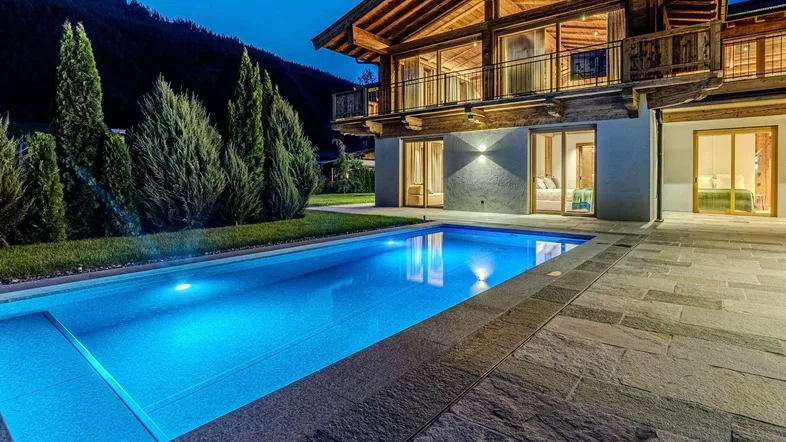 Beleuchteter Außenpool mit angrenzender Steinterrasse und luxuriösem Chalet im alpinen Stil bei Nacht, umgeben von Bäumen und Bergen.