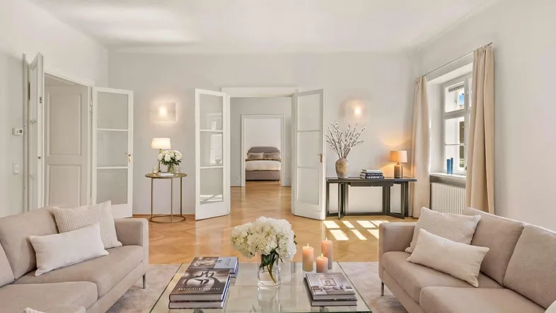 Helles Wohnzimmer mit eleganten Sofas, Parkettboden und Blick in ein Schlafzimmer.