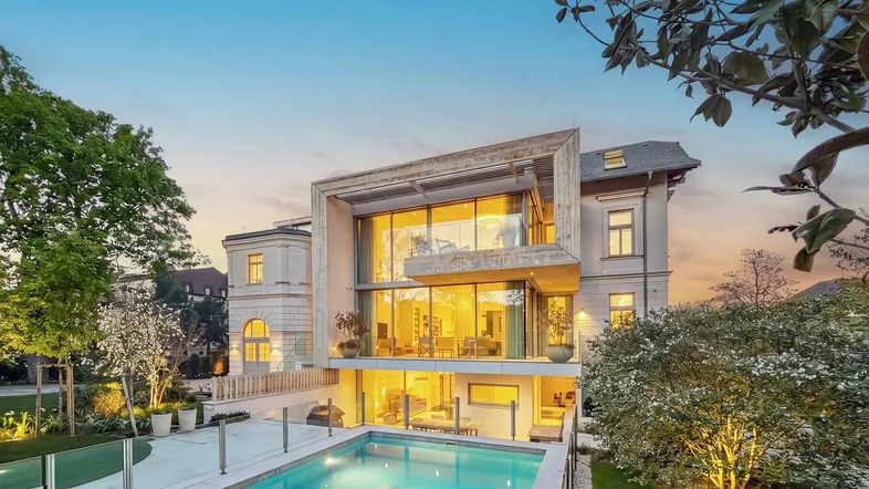 Luxuriöse Villa mit modernem Anbau, großem Garten und beleuchtetem Pool bei Sonnenuntergang.