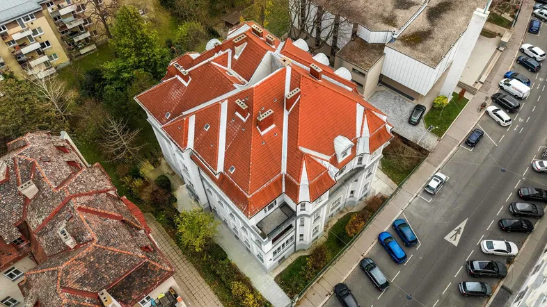 Luftaufnahme eines historischen Gebäudes mit markantem roten Ziegeldach und umliegender Stadtlandschaft.