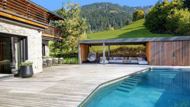Außenbereich mit großem Pool, Holzdeck und überdachter Lounge-Zone vor Bergkulisse.
