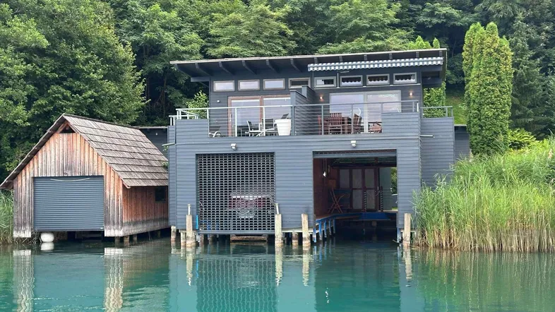 Modernes Haus am Wasser mit Bootsanleger und angrenzendem Holzschuppen, umgeben von Bäumen.
