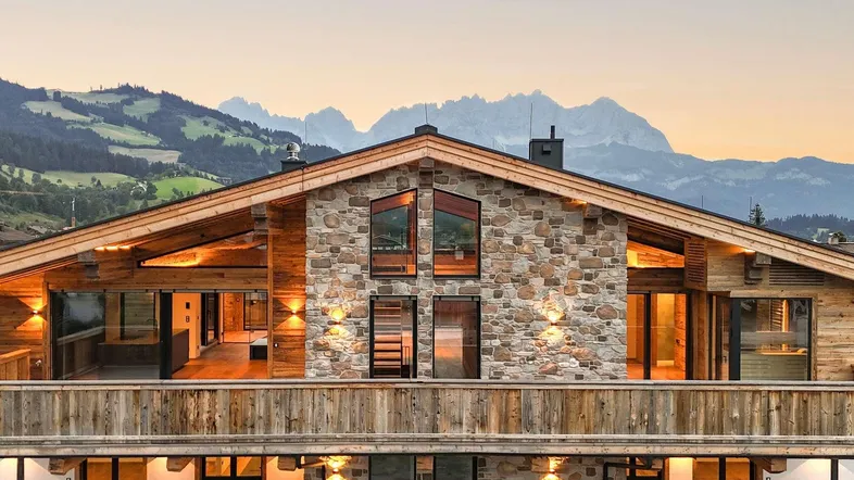 Beeindruckendes Chalet mit Stein- und Holzfassade vor einer malerischen Bergkulisse bei Sonnenuntergang.