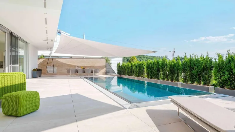 Moderne Poolanlage mit großem Schwimmbecken, Sonnenterrasse und grüner Hecke.