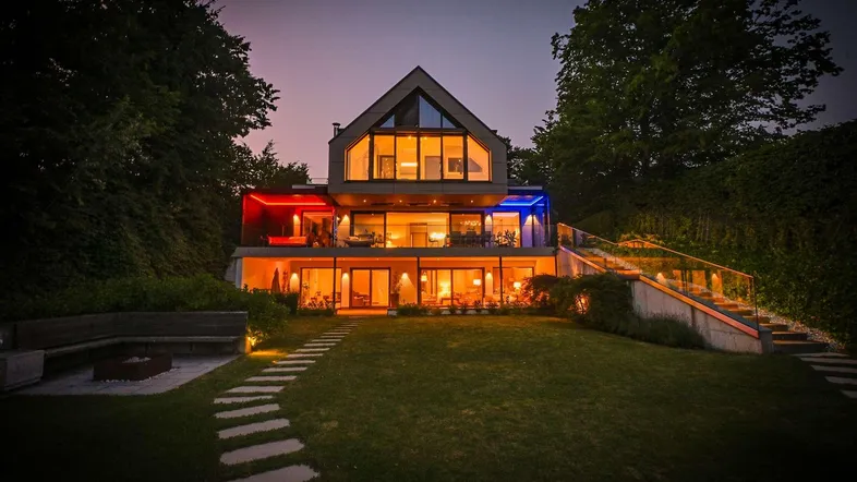 Modernes Haus bei Dämmerung mit beleuchteter Fassade, Garten und Zugang zum See.