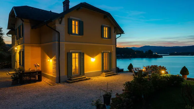 Elegantes Haus am Seeufer bei Sonnenuntergang mit beleuchteter Fassade und Garten.