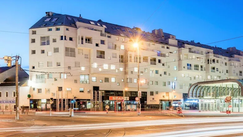 Modernes Gebäude an einer belebten Straßenkreuzung bei Dämmerung mit hellen Lichtern und Geschäften.