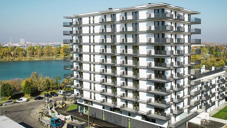 Modernes Hochhaus mit vielen Balkonen und Blick auf einen See, umgeben von Bäumen und Stadtlandschaft.