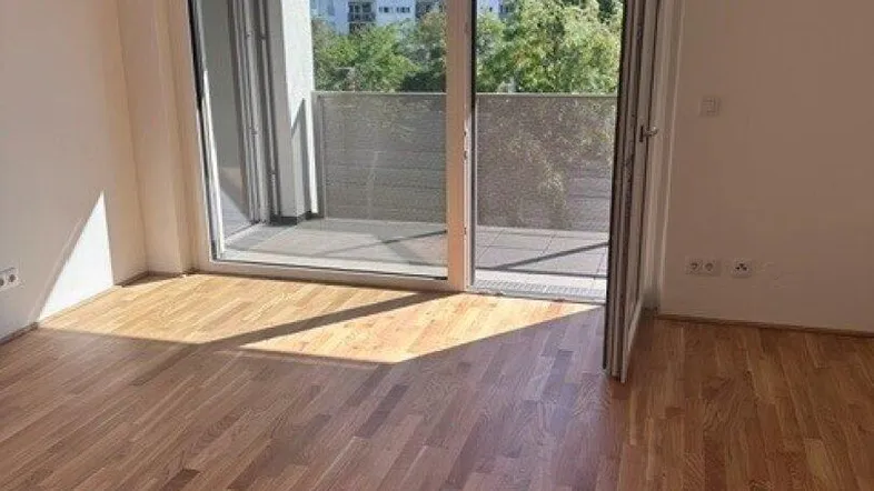 Helles Zimmer mit Parkettboden und Zugang zu einem Balkon, bietet Blick auf die umliegenden Gebäude.