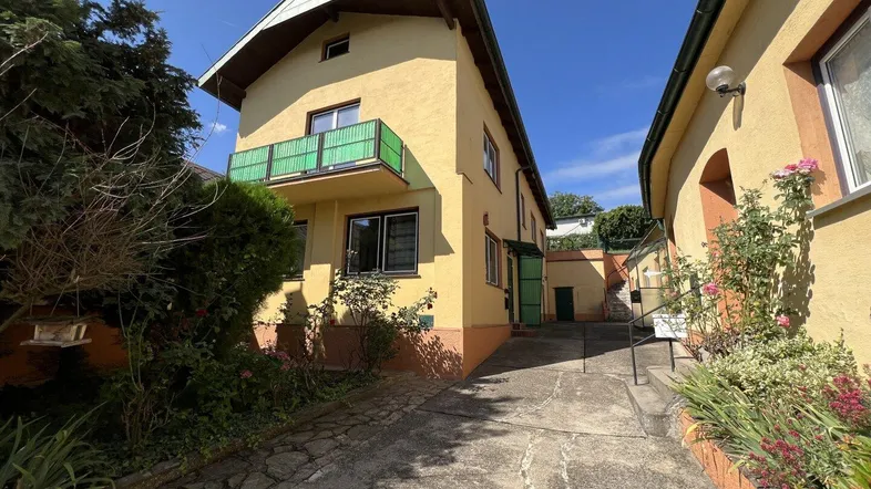Gelbes Haus mit grünem Balkon und Zufahrt, umgeben von üppiger Vegetation unter blauem Himmel.