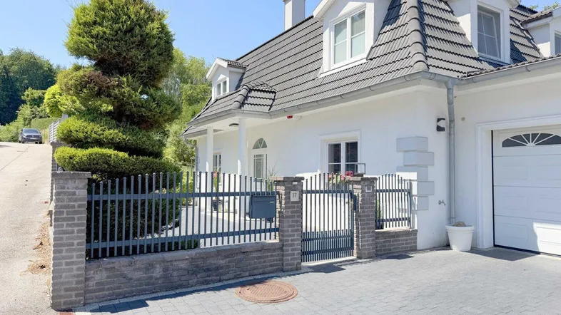Weißes Einfamilienhaus mit dunklem Ziegeldach, Vorgarten und gepflasterter Einfahrt.