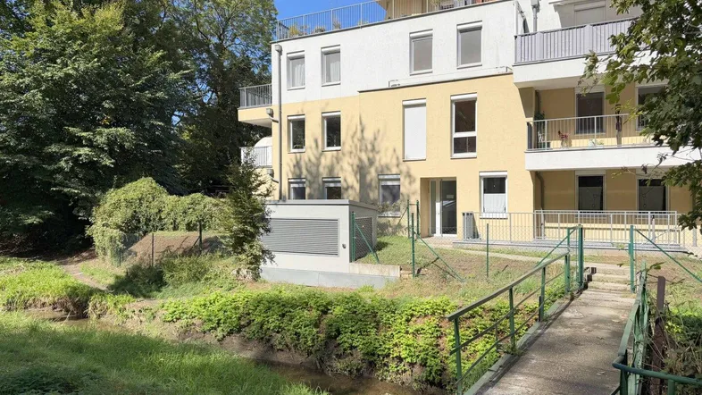 Modernes Mehrfamilienhaus mit Balkonen und Blick auf einen kleinen Bachlauf und viel Grün.