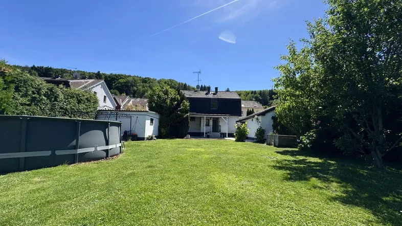 Großer Garten mit Rasenfläche und einem Aufstellpool, umgeben von Bäumen und einem Wohnhaus im Hintergrund.