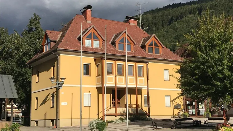 Ansicht des gelben Gebäudes mit rotem Dach und Gaubenfenstern vor einer bewaldeten Bergkulisse.