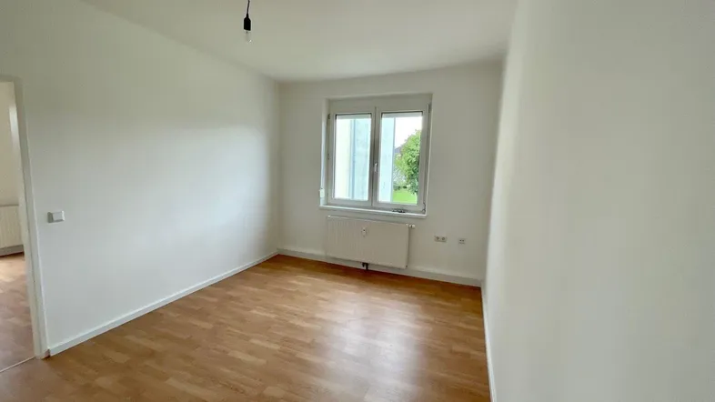 Helles, leeres Zimmer mit Laminatboden, weißen Wänden und Fenster mit Blick ins Grüne.