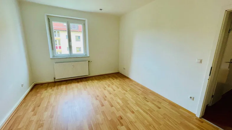 Helles Zimmer mit Laminatboden, weißen Wänden und Blick auf ein gegenüberliegendes Gebäude.