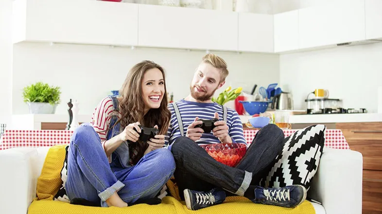 Fröhliches Paar spielt Videospiele auf einem Sofa in einem modernen, offenen Wohnbereich mit integrierter Küche.