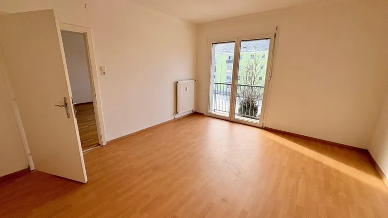 Helles Zimmer mit Holzboden, weiß gestrichenen Wänden und Zugang zu einem kleinen Balkon.