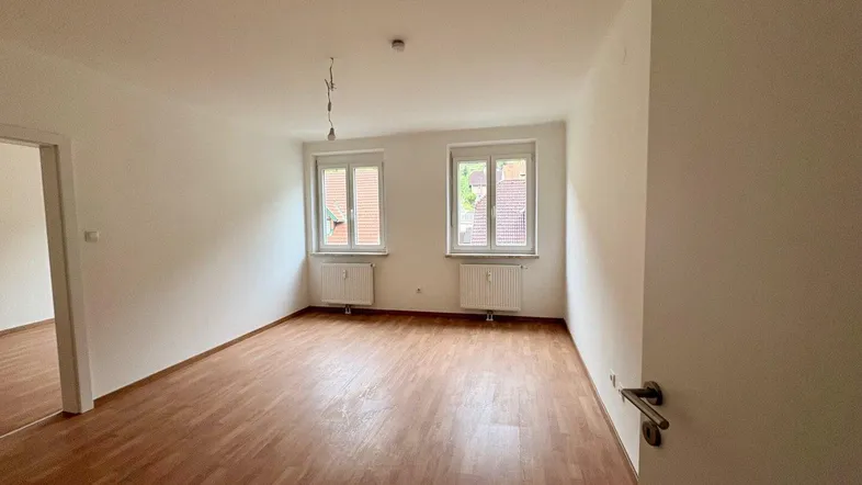 Helles, leeres Zimmer mit Laminatboden und zwei Fenstern, bereit zur individuellen Gestaltung.