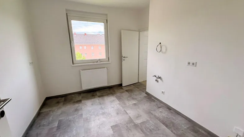 Leerer Raum mit grauem Vinylboden, Fenster und Anschlüssen für Haushaltsgeräte, ideal als Hauswirtschaftsraum.
