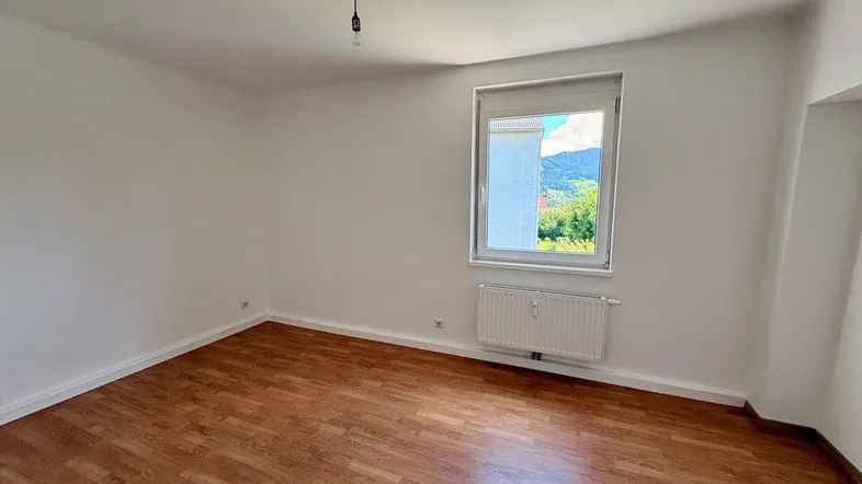 Helles Zimmer mit Laminatboden, weißen Wänden und einem Fenster mit Blick auf grüne Hügel.