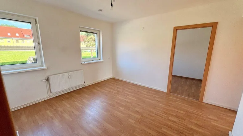 Helles Zimmer mit Laminatboden, weißen Wänden und Blick ins Grüne durch zwei Fenster.